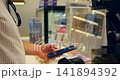 Person using smartphone for mobile payment at store checkout, hands holding phone near card reader and receipt display on payment terminal in retail setting, ready for contactless transaction 141894392