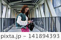 Youthful influencer browsing. Young influencer in vibrant clothing navigating online content. Young girl sporting bright attire explores digital posts along modern architectural surroundings 141894393