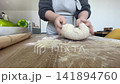 The hands of a female cook knead a lump of dough on a wooden table with white wheat flour. 141894760