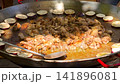 Close-up shot of chopped chicken meat browning in oil with round eggplant slices arranged along the rim of a large paella pan during an outdoor Spanish cooking festival. 141896081