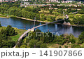 Drone view of Herrenkrug pedestrian bridge crossing Elbe River in Magdeburg, surrounded by green parkland and urban scenery. Summer cityscape shows waterfront, trees, architecture and transport links 141907866