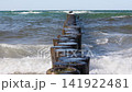 Stormy Baltic waves crash over wooden groynes near sandy shore under pale summer sky. Dynamic Ostsee seascape captures coastal energy, rough water, sea foam, windy mood and fresh seaside atmosphere 141922481