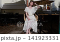 Young woman seated on wicker chair at sunlit outdoor cafe terrace, wearing flowing white lace dress and light sandals, raising hand to forehead while tilting head back 141923331