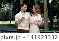 Man and woman standing on shaded park walkway holding clear cups with straws while talking, dressed in cream polo shirt and white lace dress beside trees and streetlamp 141923332