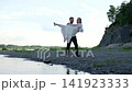 Man carrying smiling woman in flowing white dress along rocky riverbank beside calm water, with green shoreline, pale sky, and graceful outdoor wedding style mood 141923333