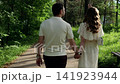 Young adults holding hands while walking along shaded forest path with soft sunlight filtering through mixed woodland, viewed from behind at eye level with calm natural mood 141923944