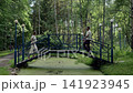 Two adults standing face to face on blue metal pedestrian bridge above algae covered pond in birch woodland with soft diffuse daylight and calm natural setting, with bridge, pond, algae, forest detail 141923945