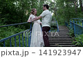 Man and woman standing close on weathered wooden stairway with blue railings in lush forest, sharing gentle touch, steady gaze, and brief kiss amid soft daylight 141923947