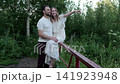 Woman and man on weathered red railing in green woodland meadow, seated pose with supportive touch, arms wrapped around shoulders, soft daylight and calm natural setting 141923948