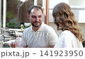 man and woman standing together, interacting in casual outdoor cafe setting with wooden furniture and drinks, capturing upper bodies of subjects in medium shot. 141923950