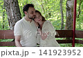 Affectionate pair resting on wooden bench inside shaded forest gazebo, woman leaning head on man's shoulder while arm wraps waist and he looks upward in calm outdoor moment 141923952
