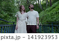 Two people standing on weathered blue footbridge in dense green forest, one resting head on shoulder while both gaze upward toward leafy canopy in calm outdoor moment 141923953