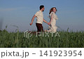 Young man and woman walking hand in hand through sunlit grassy meadow under clear blue sky, smiling with relaxed movement and natural outdoor freedom, with handholding, walking, romance, together 141923954