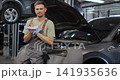 Young auto mechanic standing at open car and repairing motor in big garage. Man in uniform working in automobile repairment and smiling cheerfully to camera. 141935636