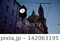 A lit street clock at dusk with the green dome and bell tower of St. Nicholas Church behind, Lesser Town, Prague, Czech Republic 142063195