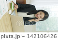 A young Japanese businesswoman is using a calculator while talking on her smartphone by a bright window (vertical video). 142106426