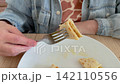 Person holding fork with piece of thin pancake over white plate on wooden table. Homemade breakfast, crepe dessert, sweet snack, simple meal. 142110556