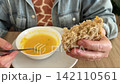 Person holding piece of whole grain bread and spoon over bowl of pumpkin soup at wooden table. Healthy lunch, homemade meal, vegetarian food. 142110561