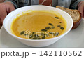 White bowl of creamy pumpkin soup with green seeds and slice of bread on plate, person at table. Healthy lunch, vegetarian meal, autumn dish. 142110562