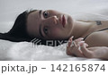 Body beauty and fashion advertisement. Female in underwear laying on the bed in the studio. Young woman seminude in lingerie laying posing under thin sheet of oilcloth wrap, staring at camera. 142165874