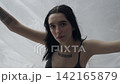 Body beauty and fashion advertisement. Female in underwear sits on the bed in the studio. Young woman seminude in lingerie sits posing under thin sheet of oilcloth wrap, looking at camera. 142165879