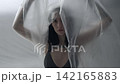 Body beauty and fashion advertisement. Female in underwear sitting on the bed in the studio. Young woman seminude in lingerie sits posing under thin sheet of oilcloth wrap, looks at camera. 142165883