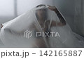 Body beauty and fashion advertisement. Female in underwear sitting on the bed in the studio. Young woman seminude in lingerie sits posing under thin sheet of oilcloth wrap, looks at camera. 142165887