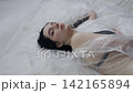 Body beauty and fashion advertisement. Female in underwear laying on the bed in the studio. Young woman seminude in lingerie laying posing under thin sheet of oilcloth wrap, looking at camera. 142165894