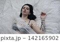 Body beauty and fashion advertisement. Female in underwear laying on the bed in the studio. Young woman seminude in lingerie laying posing under thin sheet of oilcloth wrap, looking at camera. 142165902