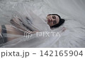 Body beauty and fashion advertisement. Female in underwear laying on the bed in the studio. Young woman seminude in lingerie laying posing under thin sheet of oilcloth wrap, looking at camera. 142165904