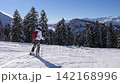 Skilled woman skiing down pristine slope on sunny winter day. Skier enjoying winter sports with beautiful mountain landscape in background. Surrounded by snow covered pine trees. Following skier 142168996