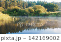 Morning landscape with mist moving over calm river surface, reflecting lush green forest on opposite bank during tranquil sunrise with golden sunlight. View from boat rafting on river 142169002