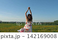 Carefree young woman wearing long floral dress spinning and dancing with her arms up in sunlit meadow of yellow dandelions. Female enjoying freedom and warm summer weather 142169008