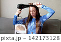 A young woman with dark blond long hair blow-dries her hair and styles her bangs with a round brush while sitting in front of a mirror at a table 142173128