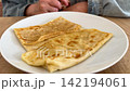 Folded thin pancakes on white plate on wooden table, person sitting behind. Homemade breakfast, crepe dessert, sweet snack, simple family meal. 142194061