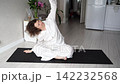 Static Shot Of Woman Performing Seated Yoga Stretch And Side Bend On Mat In Bright Home Living Room 142232568