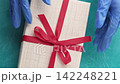 Pandemic present. Gift wrapping. Hands in protective gloves opening box untying red bow on blue textured background. 142248221
