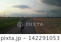 Cyclist Riding Mountain Bike Straight Ahead on Rural Asphalt Road Between Fields Towards Sunset Wind Turbines on Horizon 142291053