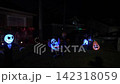 Suburban house at night with a Halloween yard full of glowing inflatables, pumpkins, and a spooky tree. Ghosts and skeleton lights decorate the lawn by parked cars. 142318059