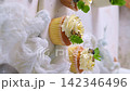 Delicious cupcakes with white frosting, nuts, blueberries, and fresh mint leaves arranged on a light textured surface with soft white fabric 142346496