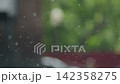 Raindrops are seen falling on a window. The background shows the green colors of trees and plants 142358275