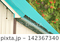 Rainwater slowly drips from the edge of a turquoise metal roof in slow motion against a blurred green garden. 142367340