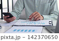 Professional financial analyst working on analyzing charts and documents using smartphone in modern office. 142370650