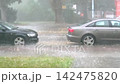 Heavy rain floods city street in Germany as parked cars stand by curb during intense downpour. Water splashes across pavement while storm reduces visibility and creates hazardous driving conditions 142475820