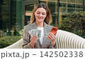 Young woman with cash and smartphone in city street celebrating business success on bench outdoors 142502338