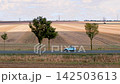Vintage retro small car drives along rural road beside harvested fields under cloudy sky. Scenic countryside scene combines classic motoring, agriculture, travel, nostalgia, and calm open landscape 142503613
