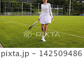 Young woman in white athletic dress bouncing tennis ball with racket on green outdoor court, female tennis player training skills before match, active lifestyle concept 142509486