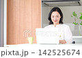 A smiling woman in her 50s typing on her computer in her kitchen at home, enjoying online work-from-home services. 142525674