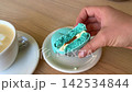 Blue Macaron Broken in Half by Female Hands Beside Cup of Coffee Showing Creamy Filling Inside French Dessert 142534844