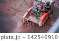 Industrial road saw cuts asphalt pavement with water cooling during construction work. Heavy cutting machine creates precise trench line while worker operates equipment on wet urban surface 142546910